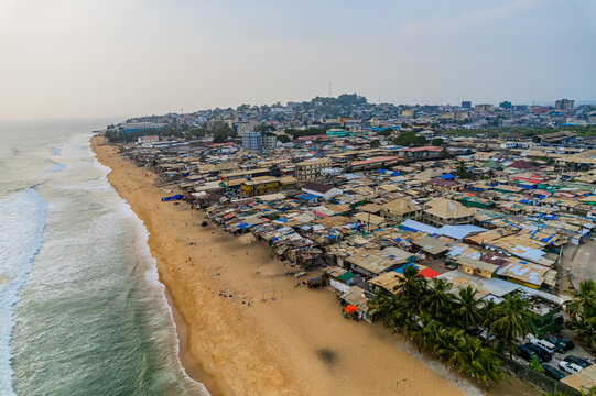 Aerial view of the dense coastal neighborhood and sandy beach with waves crashing on the shore under a hazy sky in Monrovia, Montserrado, Liberia.