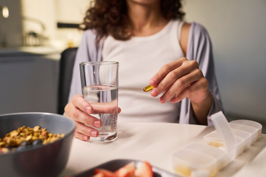 Young woman holding glass of water and taking capsule supplement at home, supporting wellness routine and influencer health content. Close up for nutrition marketing or self care campaigns