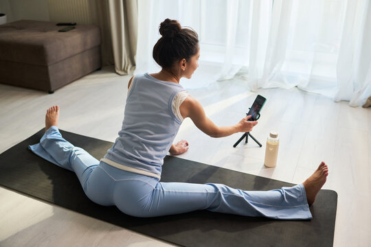 Young woman stretching on yoga mat while recording smartphone fitness content at home, showing influencer wellness routine. Ideal for social media marketing, online coaching, home workout