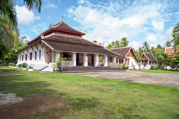 Naklejka premium Symmetrical View of Lao Buddhist Monastery Hall with Golden Door, Red Windows and Green Garden