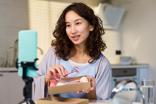 Asian young woman presenting beauty tools during livestream, holding skincare products for influencer marketing and social media promotion. Medium shot