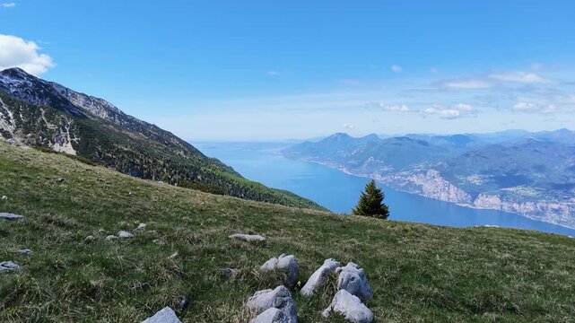 Panoramic view of Lake Garda from Monte Baldo, high altitude landscape with rocky mountain slopes, lush green meadows, blue summer sky