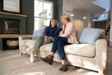 Fototapeta premium Two female partners sitting on beige sofa at home, leaning, smiling, wearing boots and sneakers