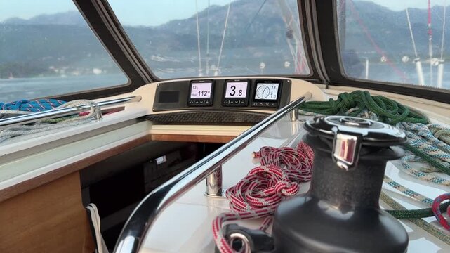 A view from inside a sailboat cockpit shows navigation instruments, ropes, and a winch, with mountains and the sea visible through the windshield