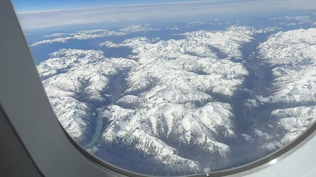 An airplane window view reveals a breathtaking expanse of snow-covered mountains and valleys under a clear blue sky, showcasing the majestic Swiss Alps near Tarasp