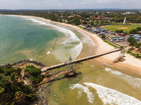 Aerial view of Parewi Duwa Temple connected by a bridge to the sandy coastline with turquoise waves and lush palm trees in Matara, Sri Lanka.