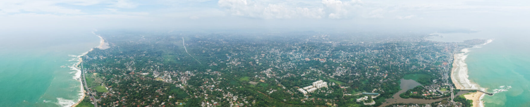 Aerial view of the sprawling coastal cityscape with dense tropical vegetation and waves crashing along the shoreline in Galle, Southern Province, Sri Lanka.