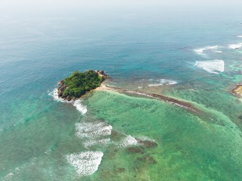 Aerial view of Devil's Rock, a small island covered in green vegetation surrounded by turquoise ocean waves and a misty horizon in Ahangama, Southern Province, Sri Lanka.