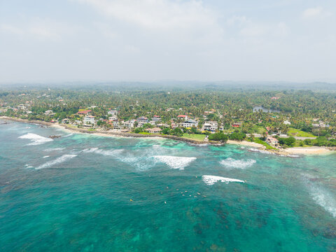 Aerial view of Devil's Rock and the turquoise coastline with surfers in the waves and lush palm trees in Ahangama, Southern Province, Sri Lanka.