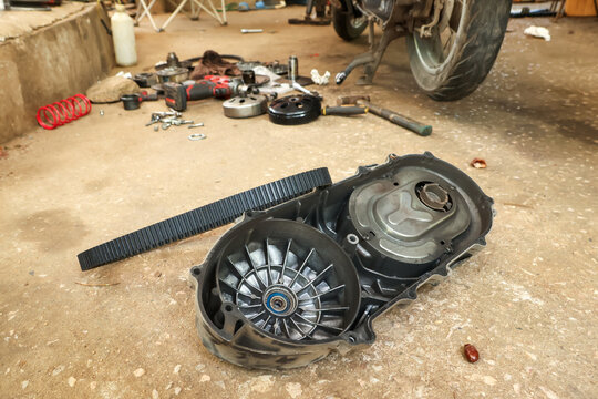 Disassembled motorcycle scooter transmission belt and clutch assembly parts resting on messy concrete garage floor conveying an industrious mechanical repair and maintenance atmosphere