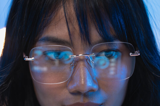 Vibrant close up of a young Asian woman software developer wearing rimless eyeglasses with bright digital programming code lines reflecting clearly on the transparent lenses