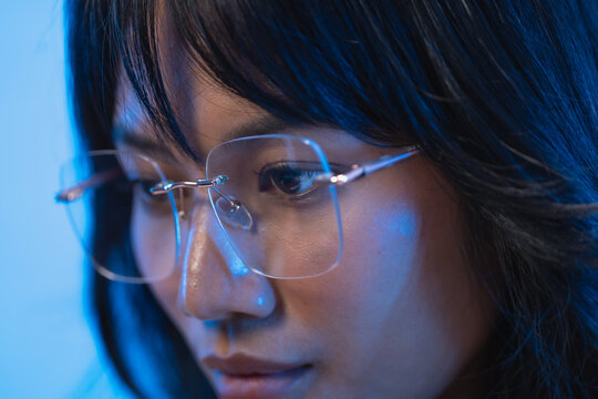 Close up profile portrait of a focused young Asian female programmer wearing transparent rimless eyeglasses while looking down at her computer screen at night with a soft cool blue
