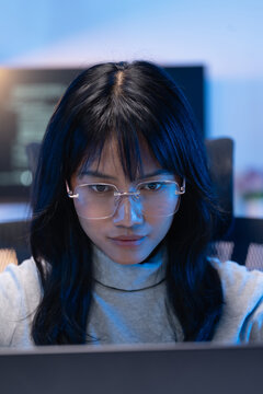 Vertical portrait of a focused young Asian female software developer wearing rimless glasses working on her laptop with a dark monitor and bright code visible behind her