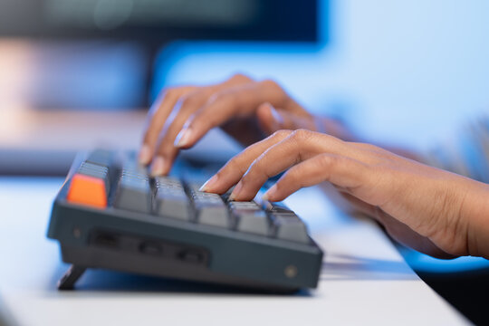 Extreme close up of young Asian woman programmer hands quickly typing program code on a black mechanical computer keyboard inside a dark blue lit modern office workspace