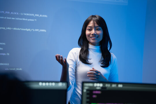 Happy young Asian female programming instructor in a white turtleneck smiling and gesturing with her hand while teaching a coding class in front of a blue digital code wall