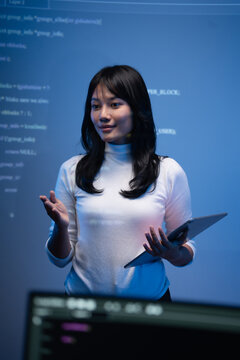 Vertical portrait of a young Asian female tech speaker in a white sweater holding a digital tablet and gesturing while explaining programming code in front of a code wall