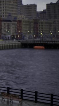 People walk along the waterfront as the sun sets. Buildings are visible in the background. An orange object floats in the water near the pier. Lights start to turn on in the buildings.