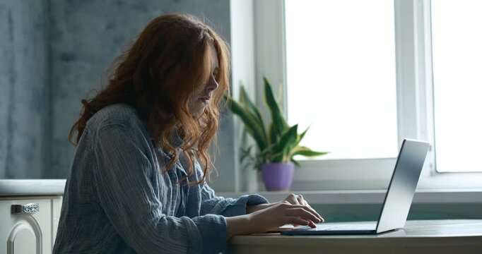 Teenage girl&rsquo;s hands typing and using the touchpad. A new modern blue computer on a table. In the background, a kitchen interior in light gray tones with a textured wall. Window light. 