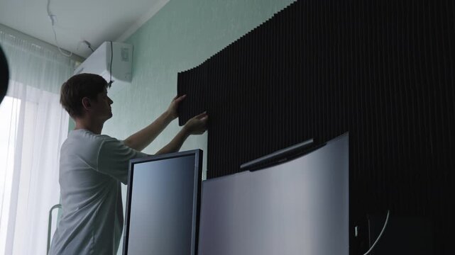 Low angle view of young man mounts black soundproof foam panels on wall of room, improving acoustics for home office or recording studio. DIY home improvement and sound absorption setup, slow motion.