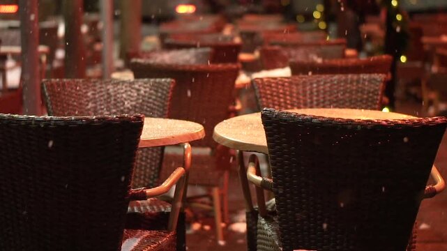 Empty tables and chairs of outdoor cafe on snowy street in evening.