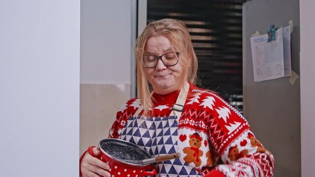 An adult woman in a festive red Christmas sweater and apron holds a cooking pot in a kitchen creating a cozy holiday lifestyle scene of winter celebration and home cooking.