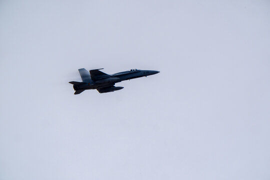Military jet aircraft in level flight against flat uniform gray overcast sky, sharp side profile with vertical stabilizer and external underwing store visible