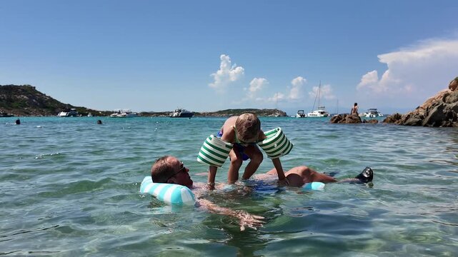 Mid-adult man and his young son wearing inflatable armbands relaxing in clear turquoise sea water on a bright sunny day during summer trip.