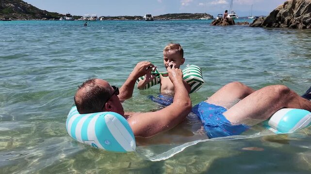 Mid-adult father and his little son in swimming goggles sitting together on an inflatable mattress in the sea, talking and laughing during holiday.