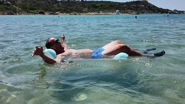 Close-up of a mid-adult man wearing sunglasses, relaxing and sunbathing on a striped inflatable mattress while floating on clear sea water waves.