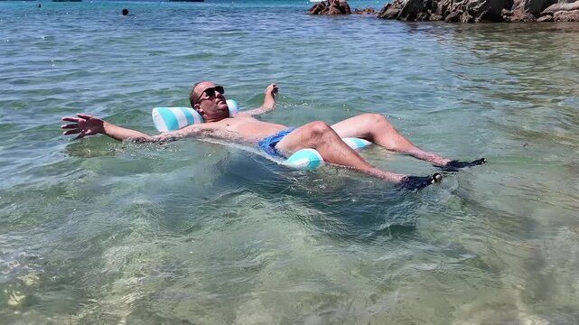 Side view of a man in swimming shorts and sunglasses floating on his back on an inflatable water mattress in a calm transparent sea.