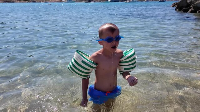 Close up shot of a little boy wearing inflatable striped armbands, successfully floating on the surface of crystal clear transparent sea water.