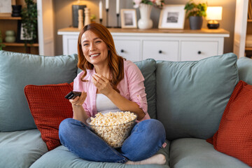 Obraz premium Woman watching TV on couch eating popcorn. Happy female sitting on sofa with remote control, home entertainment concept. Smiling girl relaxing in living room, enjoying movie and leisure time.