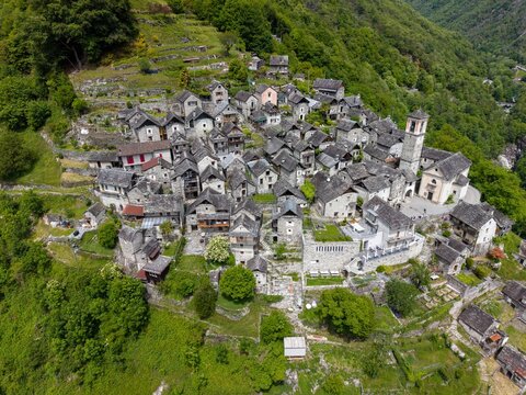 Aerial view of the historic stone houses of Corippo Albergo Diffuso nestled on a lush green hillside with traditional slate roofs and a church tower in Corippo, Ticino, Switzerland.