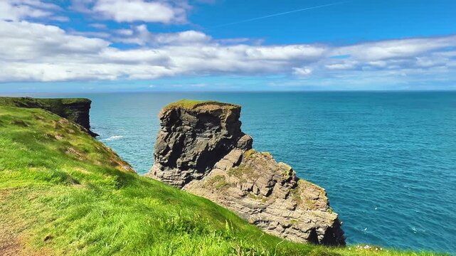 High-angle shot of a prominent sea stack and grassy cliffs meeting the ocean under a partly cloudy sky. Ideal for tourism brochures, travel agency videos, and regional marketing.