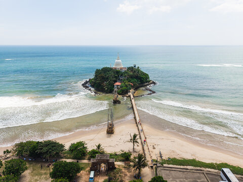 Aerial view of Parevi Duwa Temple on a small island connected by a bridge to the sandy beach with turquoise ocean waves in Matara, Southern Province, Sri Lanka.