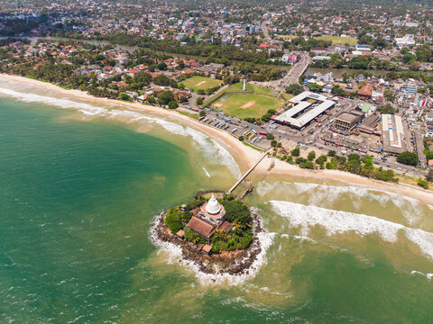 Aerial view of Parevi Duwa Temple on a small island connected by a bridge to the sandy beach and urban coastline in Matara, Southern Province, Sri Lanka.