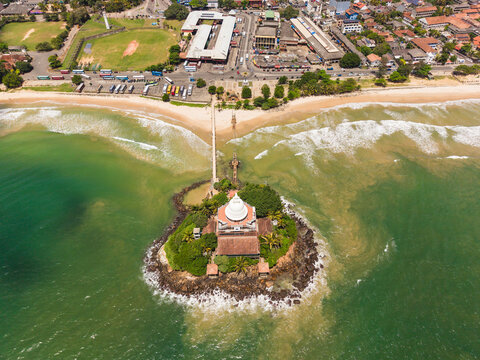 Aerial view of the Paravi Duwa Temple on a small rocky island connected by a bridge to the sandy beach and urban coastline in Matara, Southern Province, Sri Lanka.