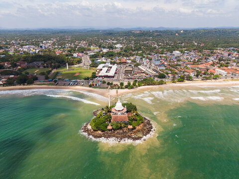 Aerial view of Parevi Duwa Temple on a rocky island surrounded by turquoise ocean waves and the coastal skyline Matara, Southern Province, Sri Lanka.