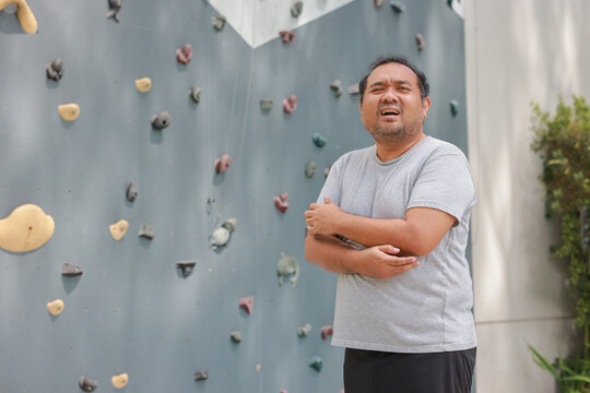 Exhausted overweight Asian man feeling arm muscle pain and holding his elbow after rock climbing workout. Sports injury, physical fatigue, and healthcare concept near outdoor bouldering wall.
