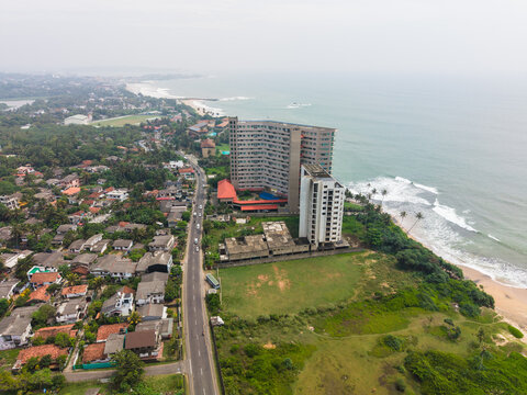Aerial view of a modern high-rise building and residential houses along the coastline with a road running parallel to the ocean in Galle, Southern Province, Sri Lanka.