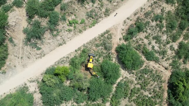 Aerial drone footage of yellow JCB backhoe loader excavator clearing dense green shrubs along a rural dirt road in dry countryside terrain with heavy construction machinery.