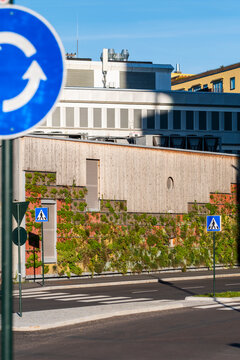 A roundabout sign frames an Oslo street with crosswalk markers, urban buildings and traffic infrastructure beneath a clear blue sky