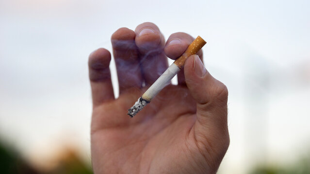 man holding burning cigarette outdoor close up view hand with smoke addiction unhealthy habit toxic tobacco nicotine substance abuse concept of healthcare, medicine, psychology