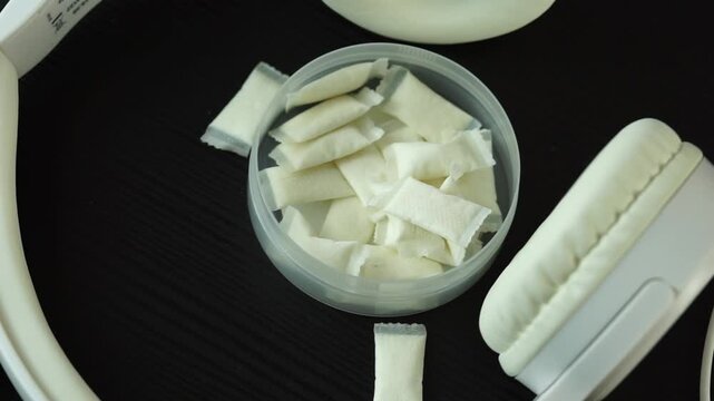 a top down view of an open round plastic container filled with small white nicotine pouches and snus pads on a dark wooden table surface surrounded by white overhead headphones