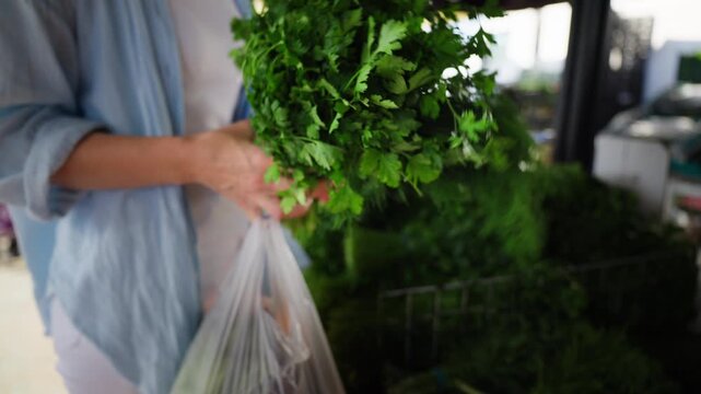 Close-up of young woman hands selecting fresh green herbs like dill and parsley at sunny farmers market, buying organic produce for healthy cooking and fresh homemade meals.
