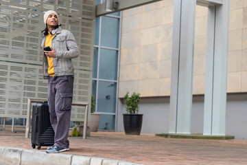 Fototapeta premium Man in his 20s standing on curb outside modern building holding smartphone and suitcase, copy space