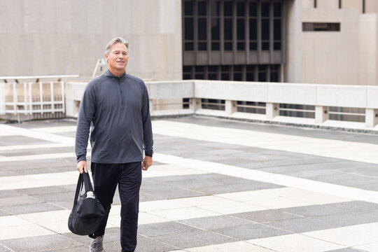 Mature adult man walking on rooftop terrace carrying black duffel bag wearing gray athletic top