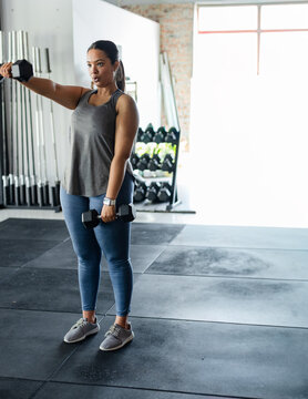 Woman holding hex dumbbells left arm extending on rubber mats in gym wearing sportswear