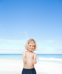 Fototapeta premium Boy standing on wide sandy beach by ocean, wearing dark swim trunks, giving thumbs up, smiling
