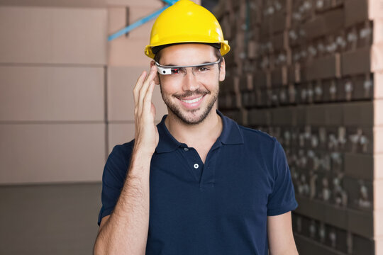 Man in navy polo wearing yellow hard hat, adjusting smart glasses in warehouse with boxes